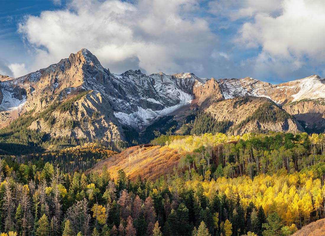 About Our Agency - View of the Mountains Against a Cloudy Blue Sky at Sunset Next to a Dense Forest with Colorful Trees