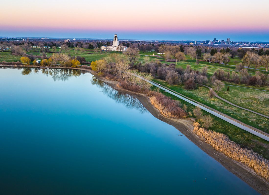 Arvada, CO - Drone View of a Lake with Surrounding Green Foliage in a Park in Arvada Colorado with Views of Downtown Denver Colorado Visible in the Background Against a Colorful Sky