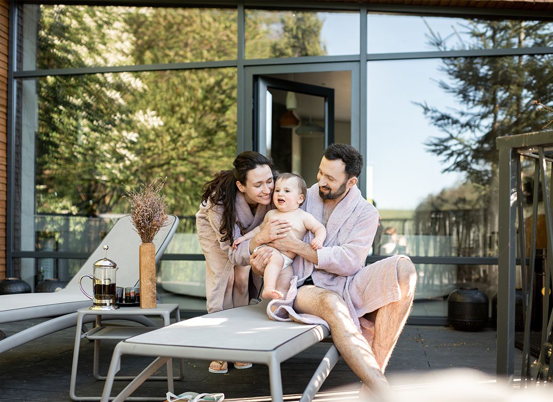 Personal Insurance - Portrait of a Cheerful Mother and Father Wearing Robes Spending Time with Their Newborn Baby on a Patio in the Backyard with Lounge Chairs